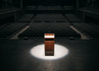 A single podium stands under a bright spotlight on a dark stage in a large hall.