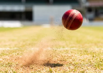 A close up view of a red cricket ball on a cricket pitch.