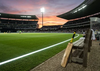A professional cricket bat leaning against a dugout bench in a stadium setting.