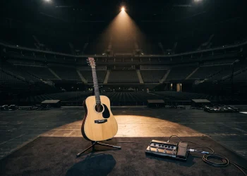 A lone acoustic guitar and loop pedal on a stage under a single spotlight in an arena.