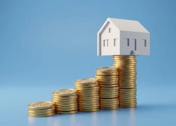 A small white house model resting on stacks of gold coins representing savings.