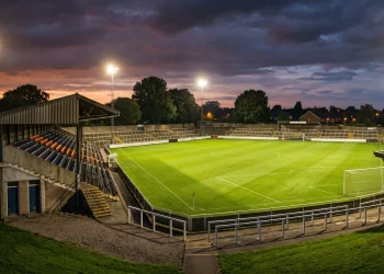 A non-league football stadium under floodlights at dusk.