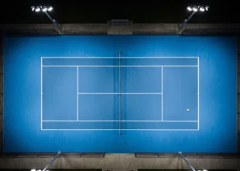 A single tennis ball on a blue hard court surface under stadium lighting.